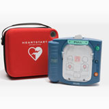 A red carrying case labeled "HeartStart" sits next to a blue Philips HeartStart AED, complete with a Philips OnSite Adult SMART Pads Cartridge and an AED pads installation guide.