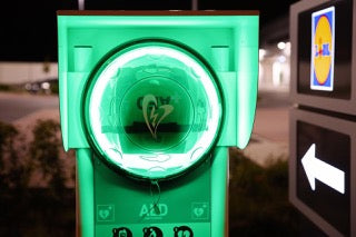 A Rotaid 24/7 AED Cabinet, equipped with full monitoring, heating, and an alarm system, illuminates in green at night as it stands ready for cardiac emergencies, with a softly lit store sign in the distance.