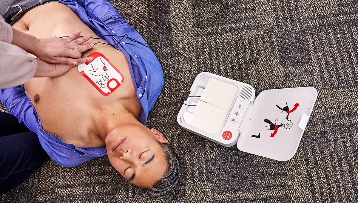 A person lies on the floor receiving CPR next to an open Physio-Control LIFEPAK CR2 USB Bilingual defibrillator by Physio-Control/STRYKER, emphasizing the crucial need for a prompt response in sudden cardiac arrest scenarios.